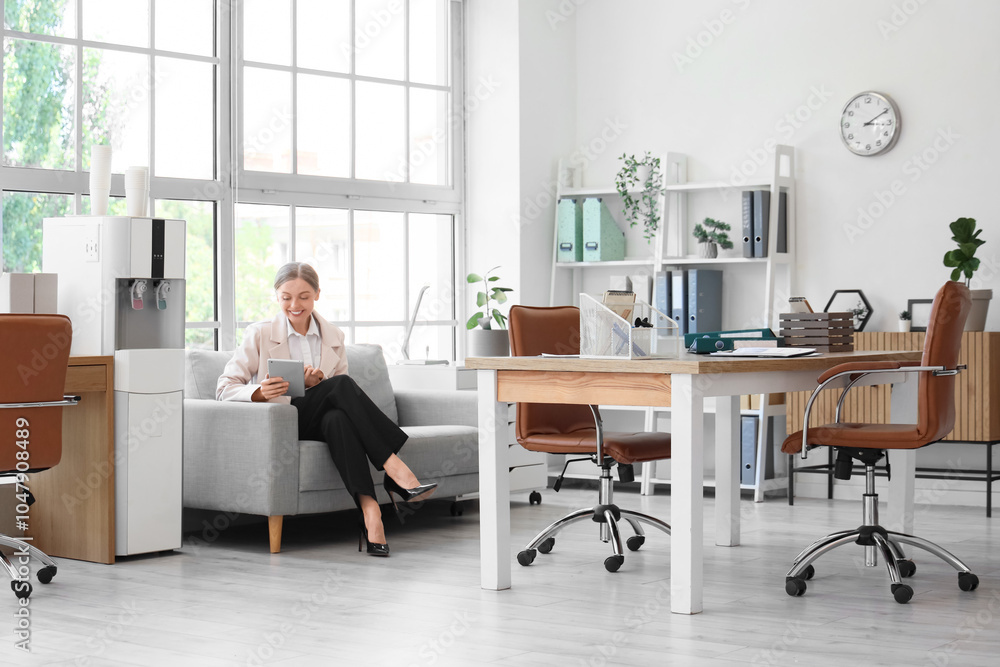 Businesswoman with tablet computer sitting on sofa near water cooler in office Businesswoman with tablet computer sitting on sofa near water cooler in office