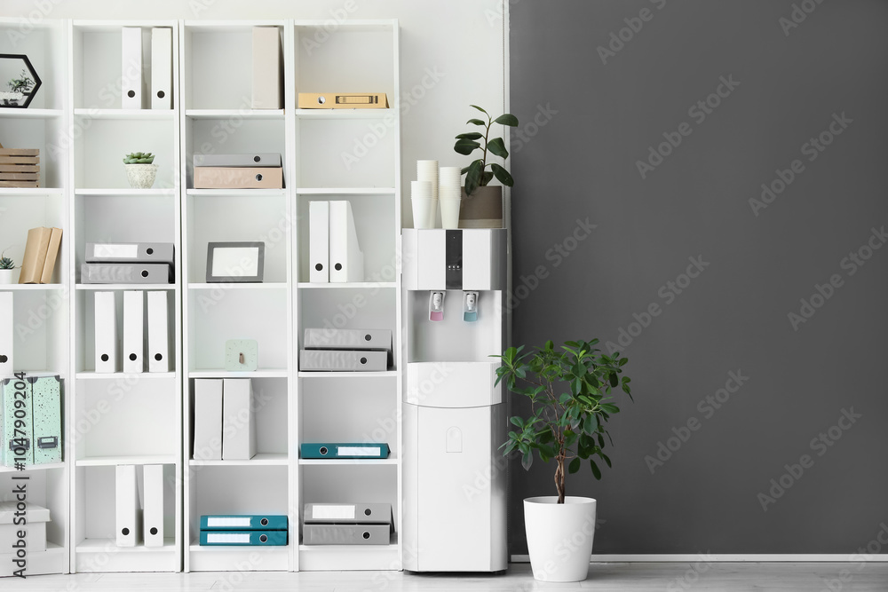 Modern water cooler with paper cups and shelf unit in office Modern water cooler with paper cups and shelf unit in office