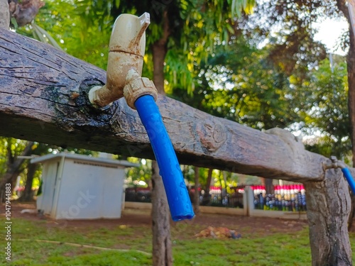 A dirty water tap attached to a wooden fence, featuring a blue hose extending from it, surrounded by lush green grass and trees, evoking a sense of outdoor utility photo