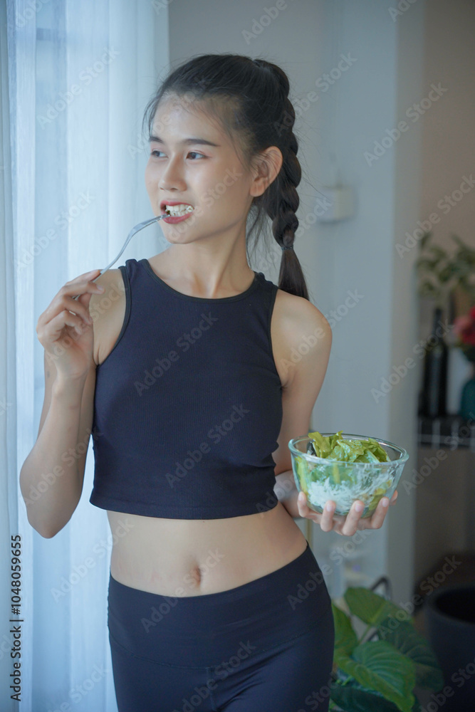 Asian woman in sportswear standing eating salad for diet in living room Asian woman in sportswear standing eating salad for diet in living room