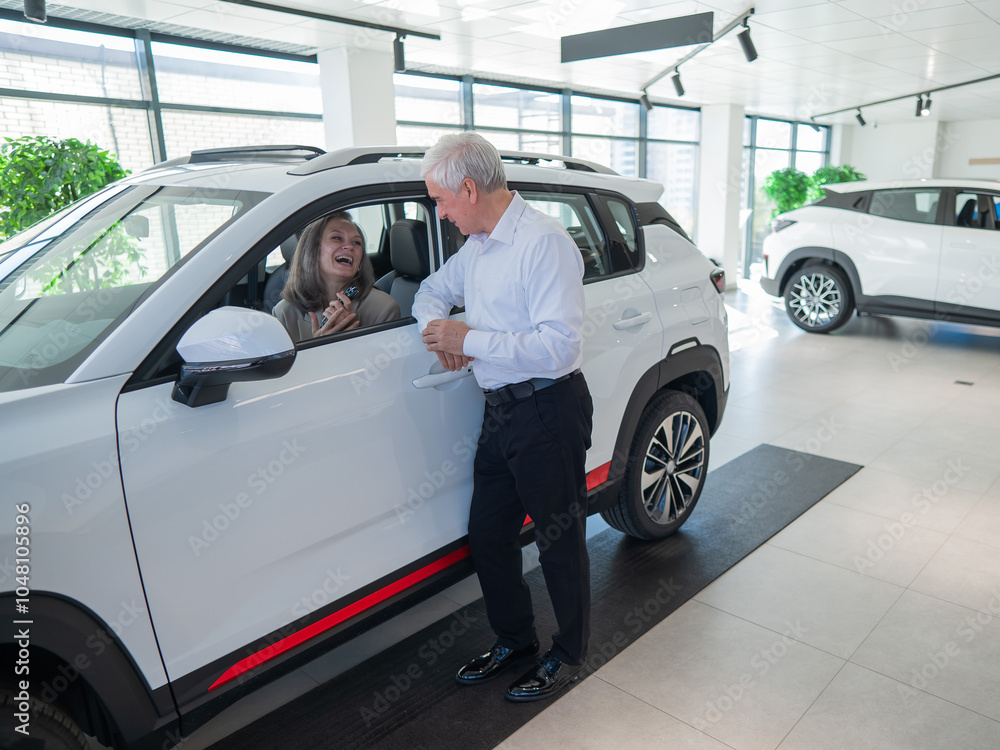 An elderly Caucasian woman is sitting in a new car. Her husband is standing nearby. An elderly Caucasian woman is sitting in a new car. Her husband is standing nearby.