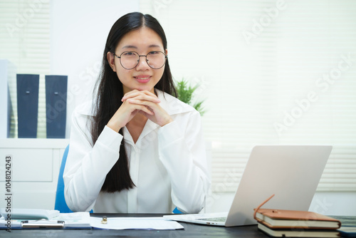 An Asian businesswoman smiles confidently while working at her desk in the office. She diligently analyzes financial data on her laptop, showcasing her expertise and commitment as an accountant