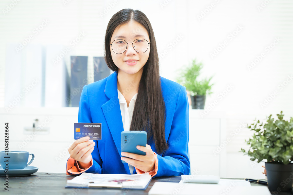 A young businesswoman sits at her office desk, smiling as she holds a credit card while shopping online. Her cheerful demeanor reflects her satisfaction with managing her work and purchases A young businesswoman sits at her office desk, smiling as she holds a credit card while shopping online. Her cheerful demeanor reflects her satisfaction with managing her work and purchases