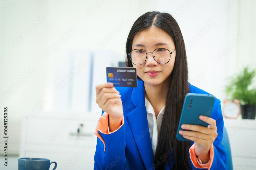 A young businesswoman sits at her office desk, smiling as she holds a credit card while shopping online. Her cheerful demeanor reflects her satisfaction with managing her work and purchases A young businesswoman sits at her office desk, smiling as she holds a credit card while shopping online. Her cheerful demeanor reflects her satisfaction with managing her work and purchases