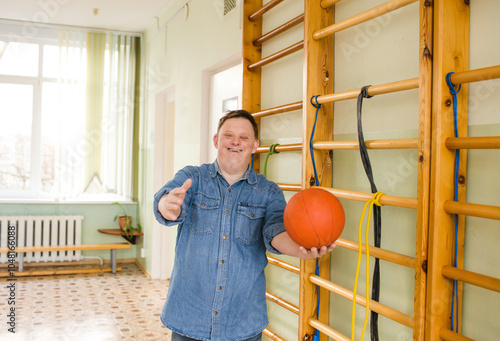 A man with Down syndrome stands in a gymnasium holding a basketball. He is smiling at the camera and looks ready to play
