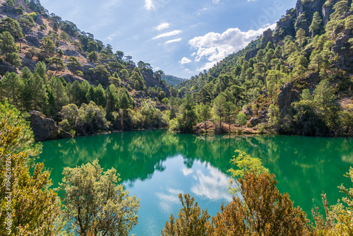 Photos Paisaje natural con un lago de aguas cristalinas que refleja un frondoso bosque y montañas bajo un cielo azul con nubes dispersas