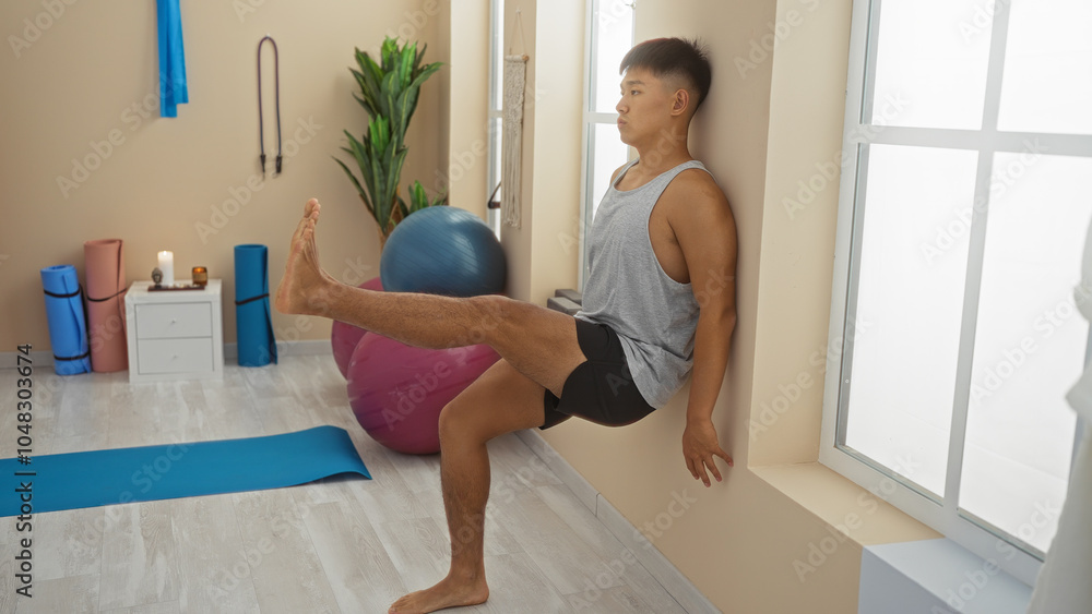 Young man performing yoga exercises in a serene spa room with yoga mats and wellness equipment, surrounded by candles and plants, wearing athletic attire, stretching against a beige wall. Young man performing yoga exercises in a serene spa room with yoga mats and wellness equipment, surrounded by candles and plants, wearing athletic attire, stretching against a beige wall.