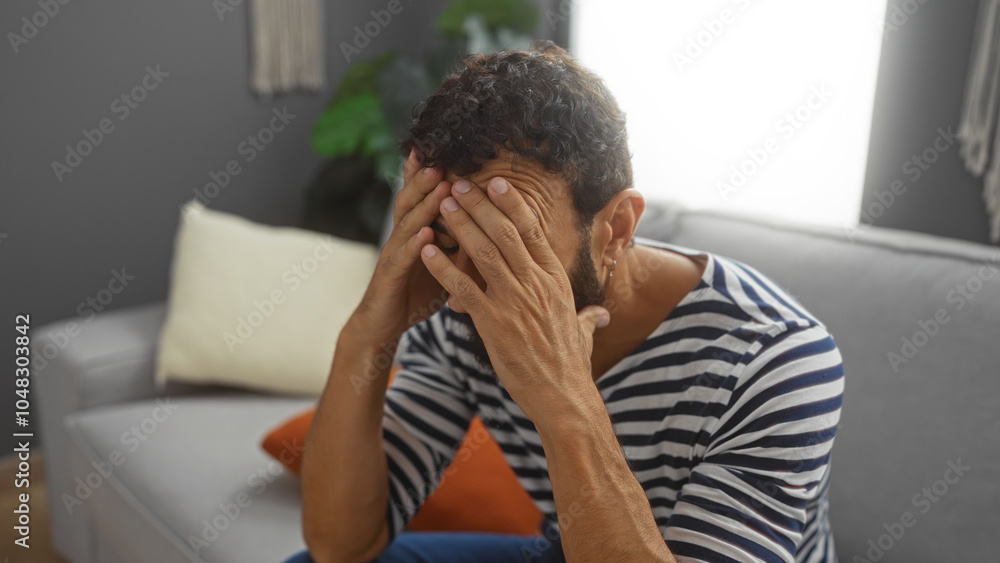 Young man with beard and striped shirt sitting on couch in living room with hands covering face looking stressed Young man with beard and striped shirt sitting on couch in living room with hands covering face looking stressed