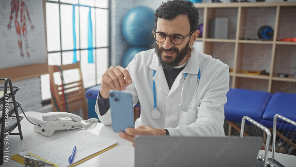 Bearded doctor using smartphone in clinic office with anatomy poster and physio balls. Bearded doctor using smartphone in clinic office with anatomy poster and physio balls.