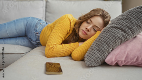 A beautiful young woman with blonde hair lies indoors on a cozy sofa in a living room wearing a yellow sweater and jeans, resting her head on a pillow with a smartphone nearby.