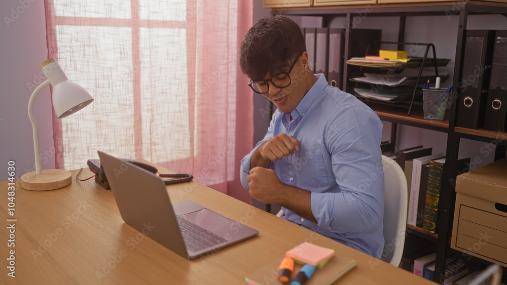 Handsome young hispanic man in an indoor office setting working at a desk with a laptop and bookshelves in the background. Handsome young hispanic man in an indoor office setting working at a desk with a laptop and bookshelves in the background.
