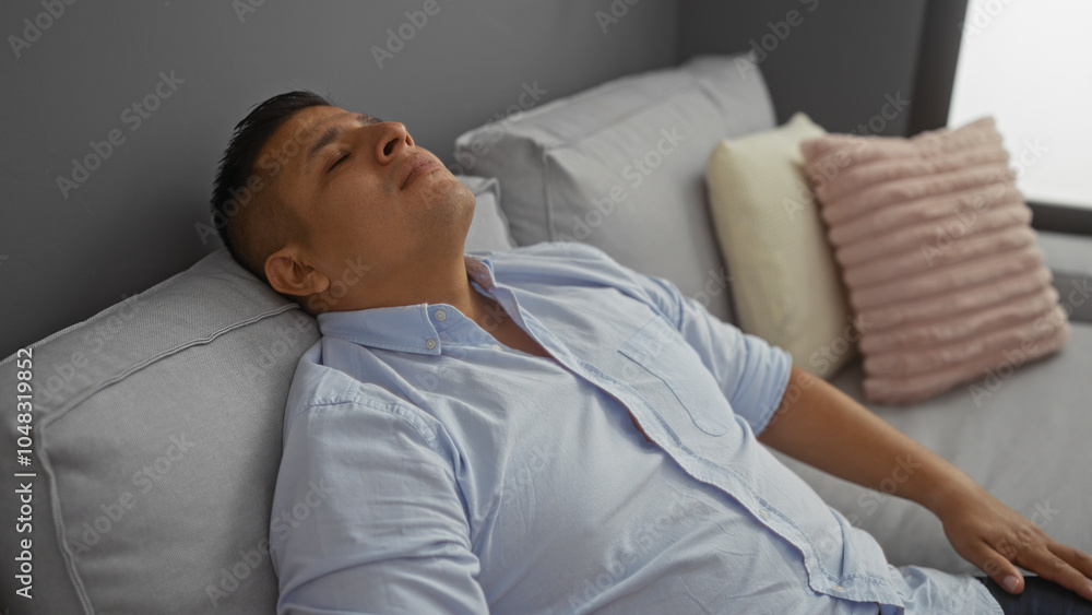 Young hispanic man relaxing on a couch in a modern living room, wearing a light blue shirt, with pillows in the background, displaying a serene expression. Young hispanic man relaxing on a couch in a modern living room, wearing a light blue shirt, with pillows in the background, displaying a serene expression.