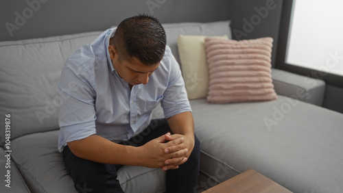 Canvas Print Young man sitting in a living room, looking down with hands clasped together, portraying a thoughtful and introspective moment in a comfortable home setting
