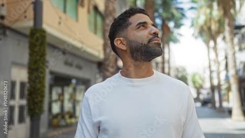 Young man looking upwards on a sunny urban street, showcasing his sharp features and stylish beard.