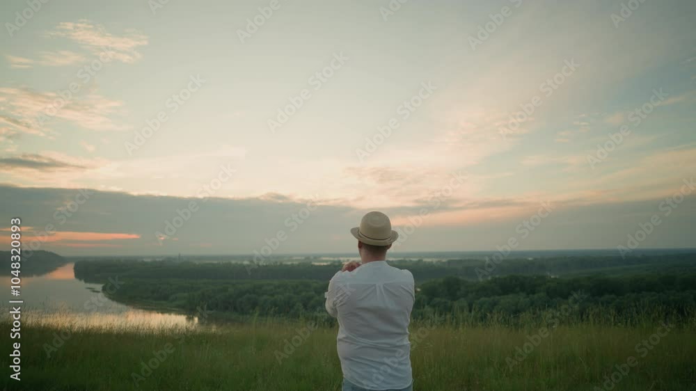 A thoughtful man wearing a white shirt, hat, and jeans stands in a grassy field by a tranquil lake at sunset, gazing into the distance. The warm sunset light create a peaceful atmosphere A thoughtful man wearing a white shirt, hat, and jeans stands in a grassy field by a tranquil lake at sunset, gazing into the distance. The warm sunset light create a peaceful atmosphere