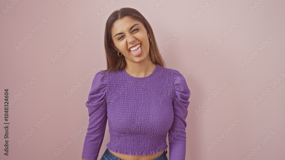 A playful young woman sticks her tongue out for a fun portrait against a pink wall, highlighting her latin ethnicity and stylish purple outfit. A playful young woman sticks her tongue out for a fun portrait against a pink wall, highlighting her latin ethnicity and stylish purple outfit.