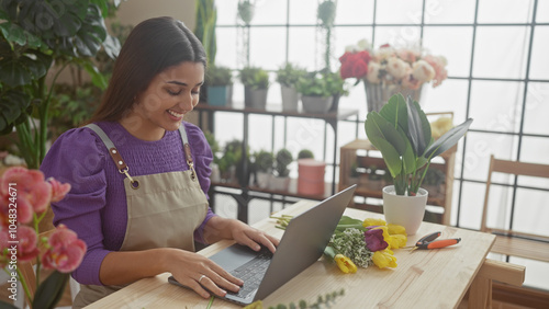 Fotografi Smiling woman using laptop in a flower shop surrounded by plants, showcasing mod