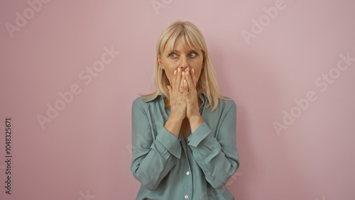 Elderly woman in teal blouse looks surprised or worried with hands covering her mouth, against an isolated pink background