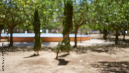 Blurred image showing an outdoor park with trees, greenery, sunlight, and defocused background featuring people near a water fountain