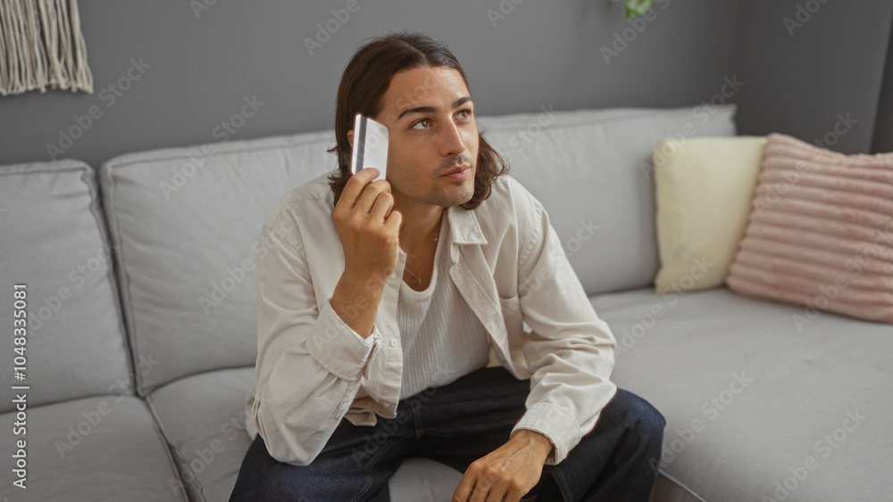 Young man with long hair sitting in a living room holding a credit card, appearing thoughtful, indoors in a home setting. Young man with long hair sitting in a living room holding a credit card, appearing thoughtful, indoors in a home setting.