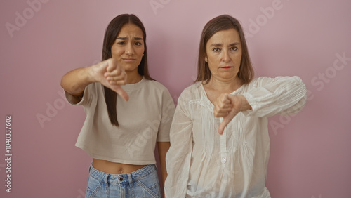 Mother and daughter showing disapproval with thumbs down against a pink wall background, conveying family dissatisfaction and negative emotions photo