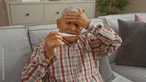 Mature hispanic man checking his temperature with a thermometer while sitting on a couch at home showing concern. photo