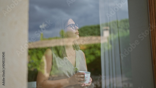 Fotografi Woman indoors looking through window with reflections holding a cup in quiet caf