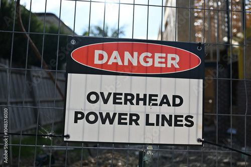 Rectangular danger overhead power lines sign attached to a tilting metal fence, outside the building site of a suburban home