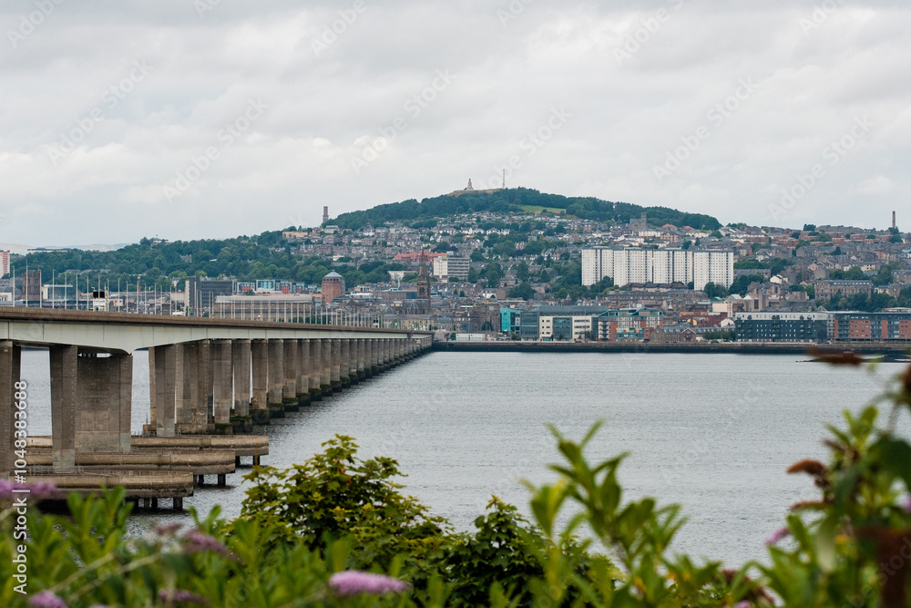 Naklejka premium Dundee Scotland: 4th Aug 2024: The Tay Bridge stretches over a calm body of water with a cloudy sky above in an urban landscape