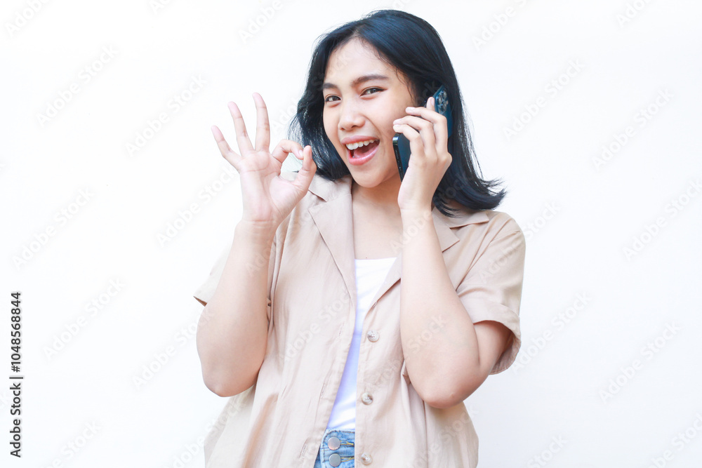 excited asian woman showing okay signs and holding smartphone wearing casual brown clothes isolated on white background excited asian woman showing okay signs and holding smartphone wearing casual brown clothes isolated on white background