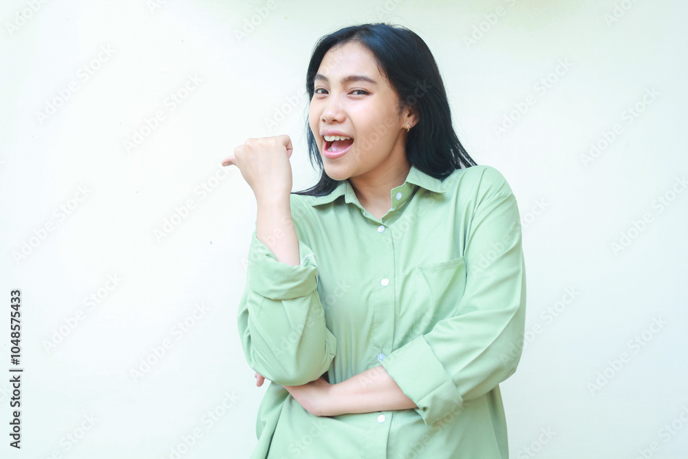 delighted success asian woman pointing to side empty space with folding arms looking at camera with mouth wide open screaming wearing green over sized clothes isolated on white background