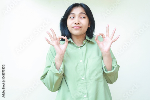 conten glad asian woman wearing green over size shirt showing ok signs with two hands and looking aside to empty space giving excellent gesture and compliment standing isolated white background photo