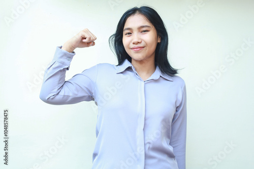 peaceful young asian business woman show strength bicep muscle with raising arm fist looking at camera with smiling relax expression, wear formal shirt, isolated on white background