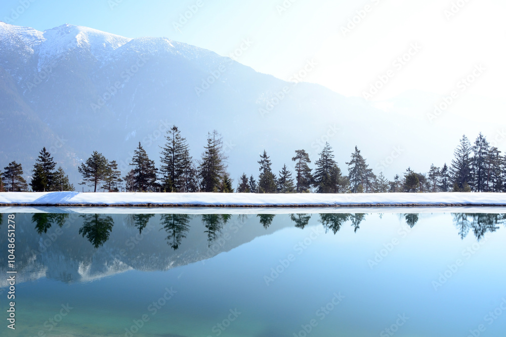 Obraz premium A panoramic view of a winter unfrozen lake in front of mountains and a row of trees. Picturesque sunny winter landscape