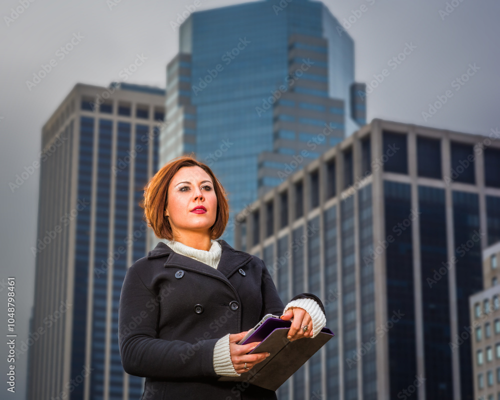 A woman stands confidently in front of towering skyscrapers, holding a tablet in her hands. The overcast sky and modern architecture create a professional and urban atmosphere..