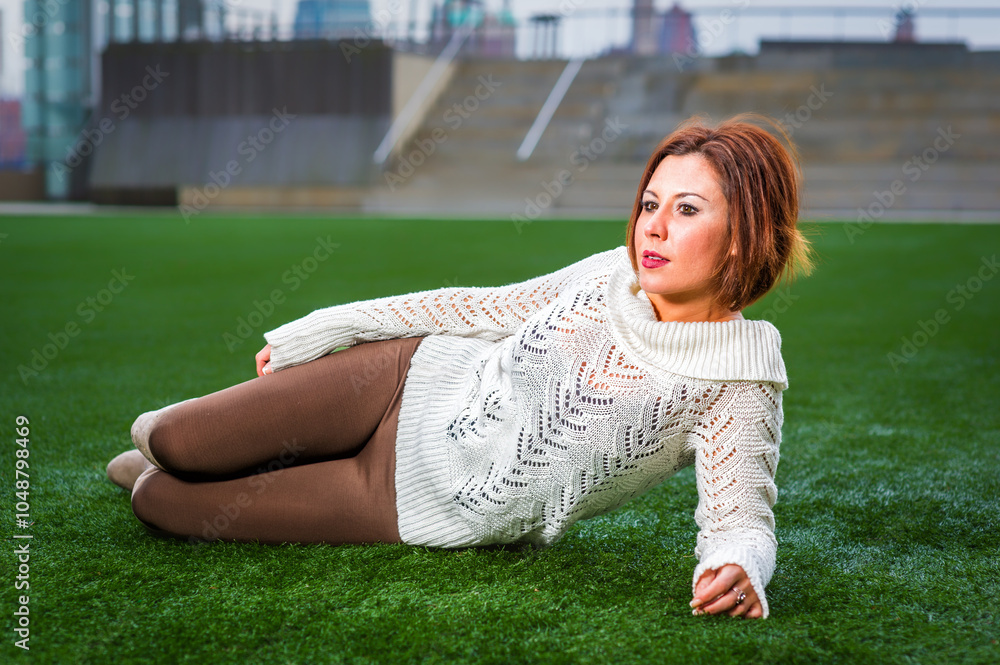 A woman relaxes on a lush green lawn, wearing a white sweater and brown pants. The setting appears to be a rooftop or outdoor area with blurred buildings in the background.