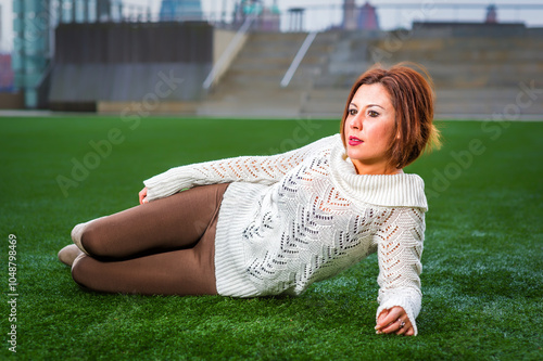 Obraz na płótnie A woman relaxes on a lush green lawn, wearing a white sweater and brown pants. The setting appears to be a rooftop or outdoor area with blurred buildings in the background. w salonie