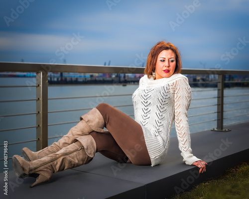 Obraz na płótnie A woman wearing a white knit sweater and brown leggings sits on a ledge with a waterfront view behind her. The setting appears to be calm and overcast, adding a serene atmosphere to the scene.. w salonie
