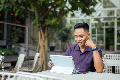 A smiling man in a purple shirt uses a tablet while sitting at an outdoor cafe table, surrounded by greenery, conveying relaxation and technology use in a casual setting.