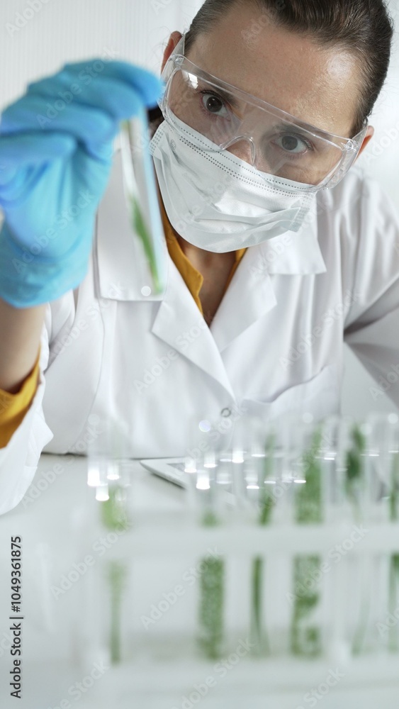 Woman scientist wearing a lab coat, blue gloves, face mask and protective glasses, is holding a test tube with plants inside, vertical portrait view. Science and medicine concept Woman scientist wearing a lab coat, blue gloves, face mask and protective glasses, is holding a test tube with plants inside, vertical portrait view. Science and medicine concept