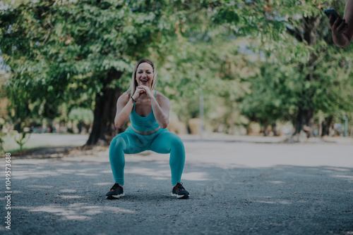 A woman in teal athletic outfit performs a squat in a scenic park, surrounded by greenery, highlighting outdoor fitness and an active lifestyle in a refreshing natural setting. photo