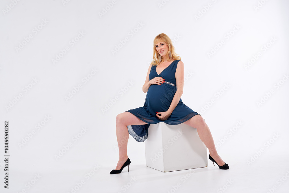 A pregnant woman is wearing a blue dress and standing in front of a white background. She is wearing black heels and has blonde hair A pregnant woman is wearing a blue dress and standing in front of a white background. She is wearing black heels and has blonde hair