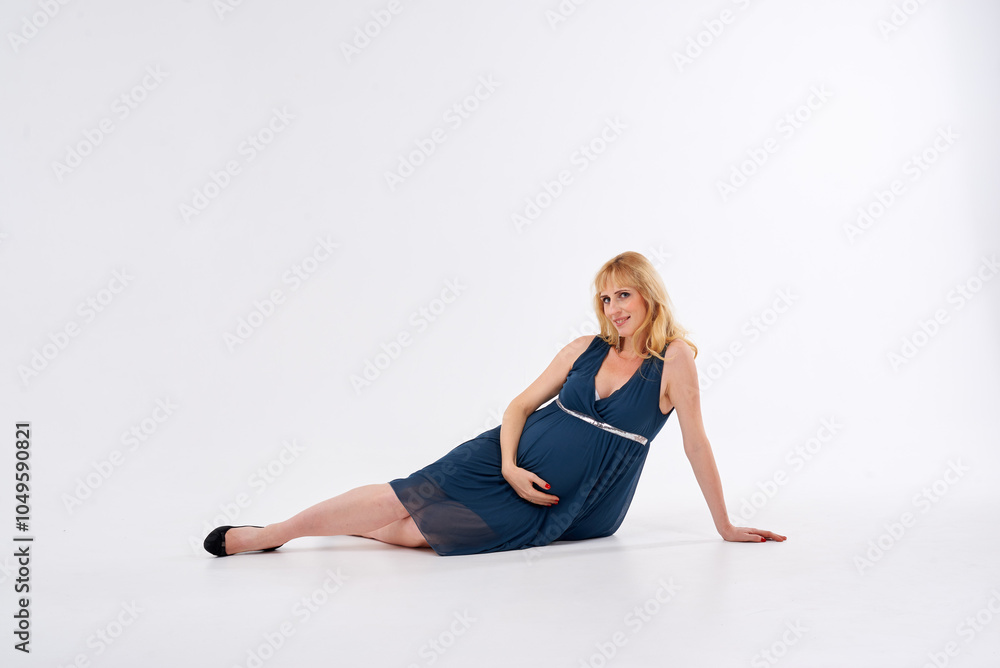 A pregnant woman is wearing a blue dress and standing in front of a white background. She is wearing black heels and has blonde hair A pregnant woman is wearing a blue dress and standing in front of a white background. She is wearing black heels and has blonde hair