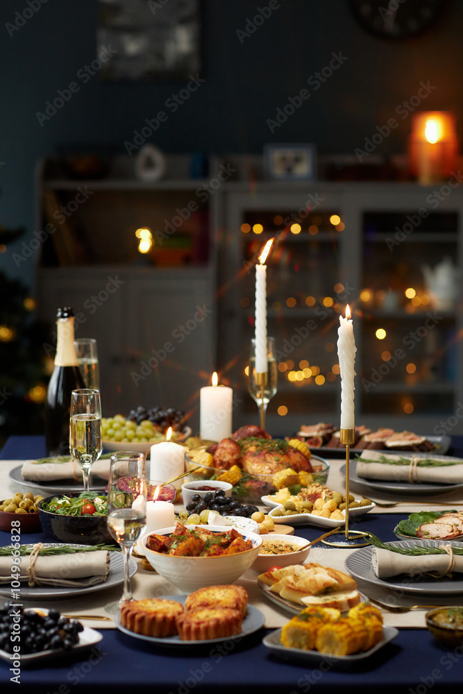 Naklejka premium Vertical shot of dining table set with variety of festive dishes, champagne glasses and lit candles while christmas lights are glowing in background, copy space