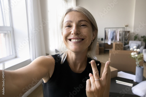 Overjoyed mature woman small retail company owner shoot selfie at work show victory v gesture to camera proud with profitable business. Smiling successful SME startupper make videocall from workplace photo