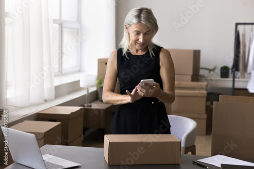 Elderly lady small company owner prepare moving to new office order courier service to transfer stuff packed in boxes. Storehouse or postal worker contact addressee by phone to check shipping details photo