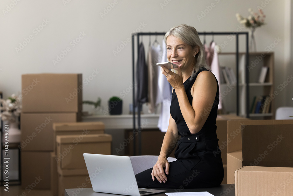 Middle age seller businesswoman working from office at web store warehouse storage using mobile phone voice app to contact customer. Self employed retail trader dictate email via speakerphone on cell