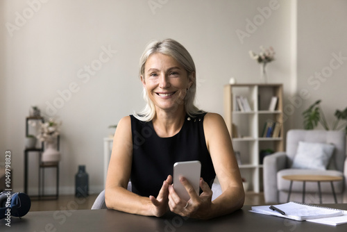 Headshot portrait smiling positive elderly female remote worker freelancer sit at home office desk use app on mobile phone. Happy aged lady look at camera enjoy using modern gadget in work study life