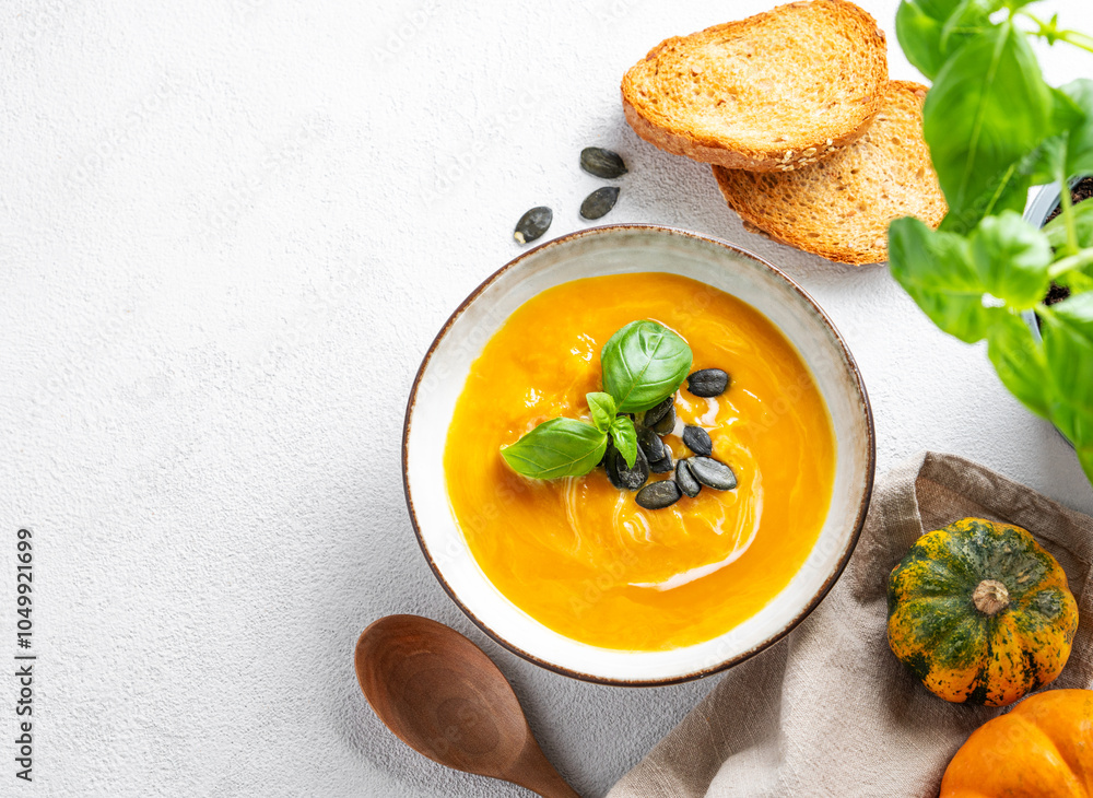 Flat lay of pumpkin soup with cream, basil and seeds on a light background with bread, spoon, fresh pumpkins and herb. Flat lay of pumpkin soup with cream, basil and seeds on a light background with bread, spoon, fresh pumpkins and herb.