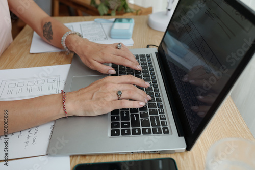 Hands engaged on laptop keyboard while designing user interface. Notes and sketches visible on desk along with digital device photo